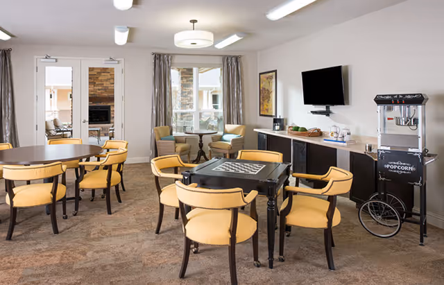 A bright common room with yellow-upholstered chairs around game and dining tables, a wall-mounted TV, popcorn machine, and seating by windows.