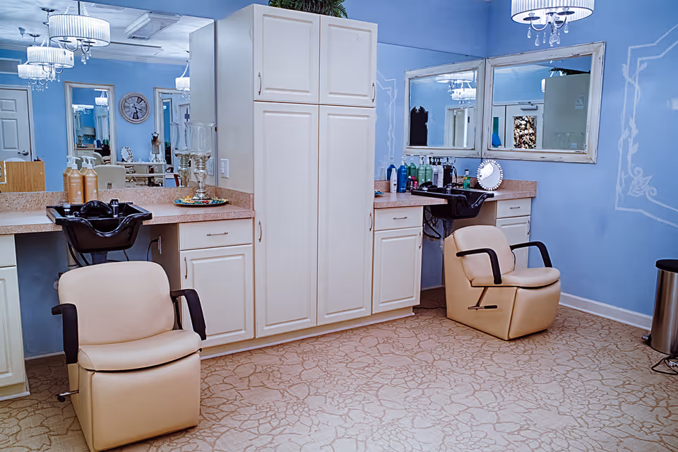 Interior of a salon area with two beige salon chairs in front of black hair washing sinks. The walls are painted light blue with white decorative trim. There are large mirrors above the countertops with various hair care products placed on them. The floor has a patterned beige carpet, and there are white cabinets and drawers for storage.