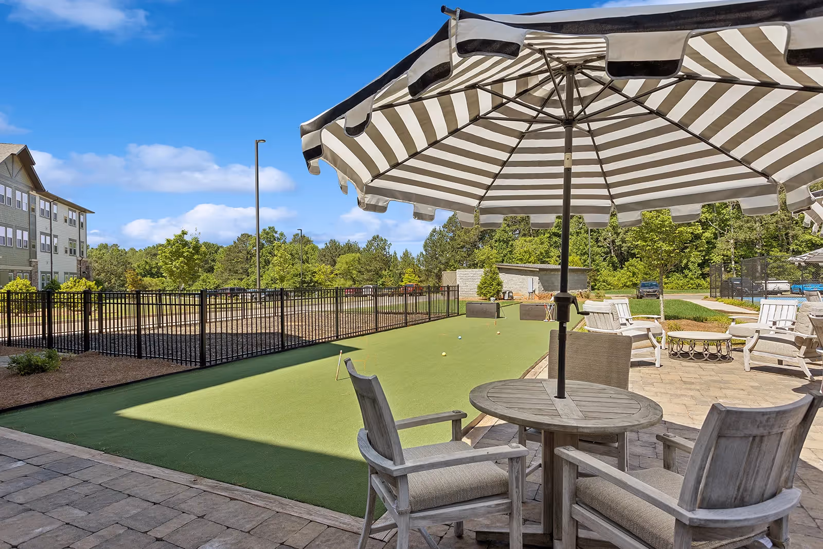 Outdoor patio area with a striped umbrella shading a round table and cushioned chairs. In the background, there is a bocce ball court surrounded by a black metal fence, additional seating with white chairs, and greenery including trees and shrubs under a blue sky with some clouds.