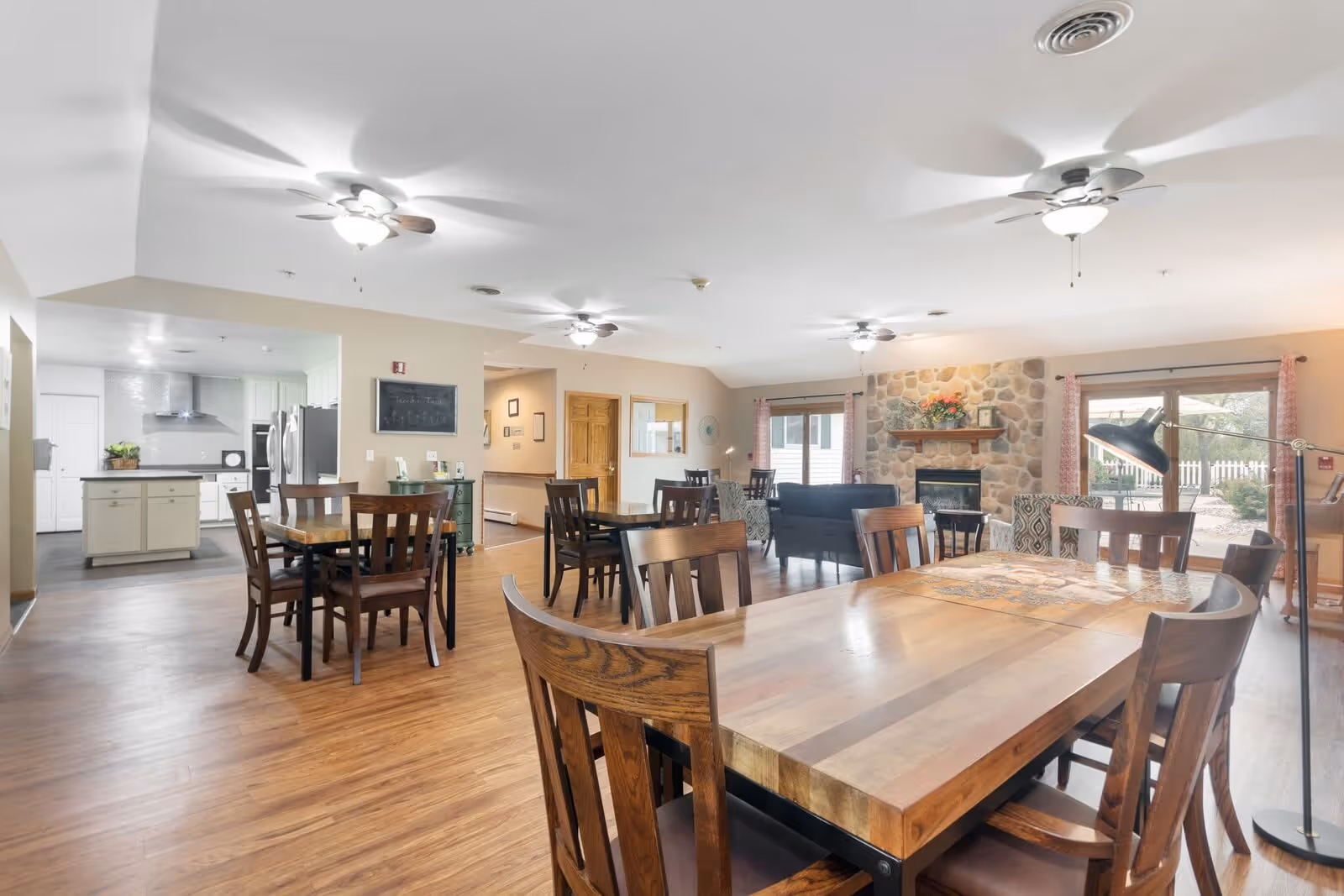A spacious and well-lit common area in an assisted living facility featuring multiple wooden dining tables and chairs, a cozy seating area with sofas near a stone fireplace, ceiling fans, and large windows with curtains allowing natural light to enter. The kitchen area is visible in the background with modern appliances and white cabinetry.