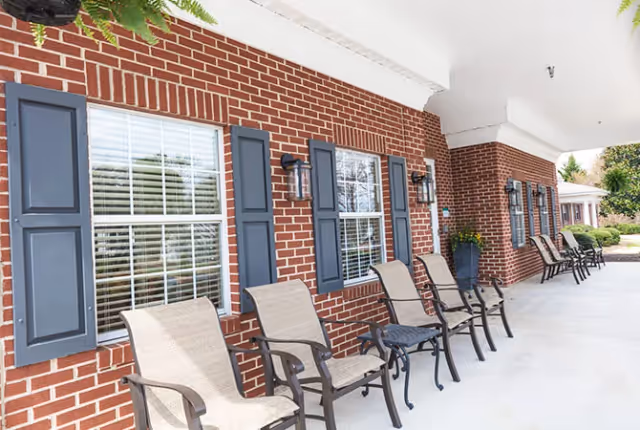 Covered outdoor patio area with a row of beige cushioned chairs and small black tables against a red brick wall with windows and blue shutters at TerraBella Athens facility.