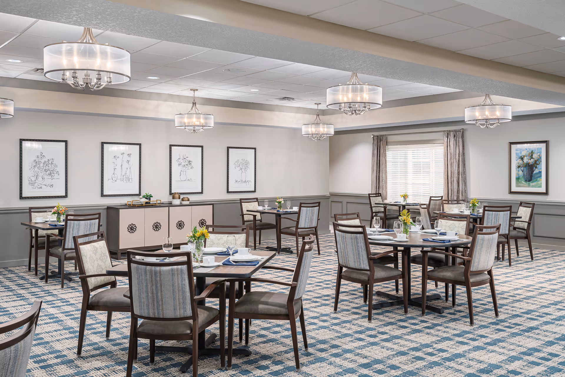 A spacious dining room with multiple tables set for meals, each table adorned with small flower arrangements. The room features patterned carpet, modern chandeliers, framed artwork on the walls, and large windows with curtains allowing natural light to enter.