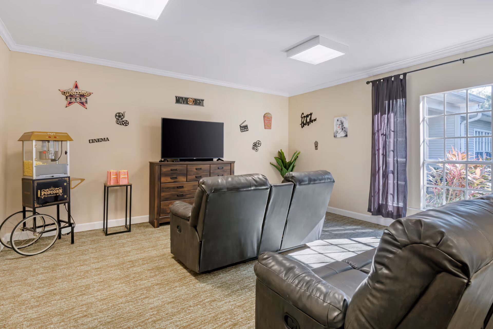 A cozy living room with two black leather recliners facing a flat-screen TV on a wooden dresser. The walls are decorated with movie-themed signs and a popcorn machine is placed on the left side. A large window with dark curtains lets in natural light, and there is a green plant in the corner.