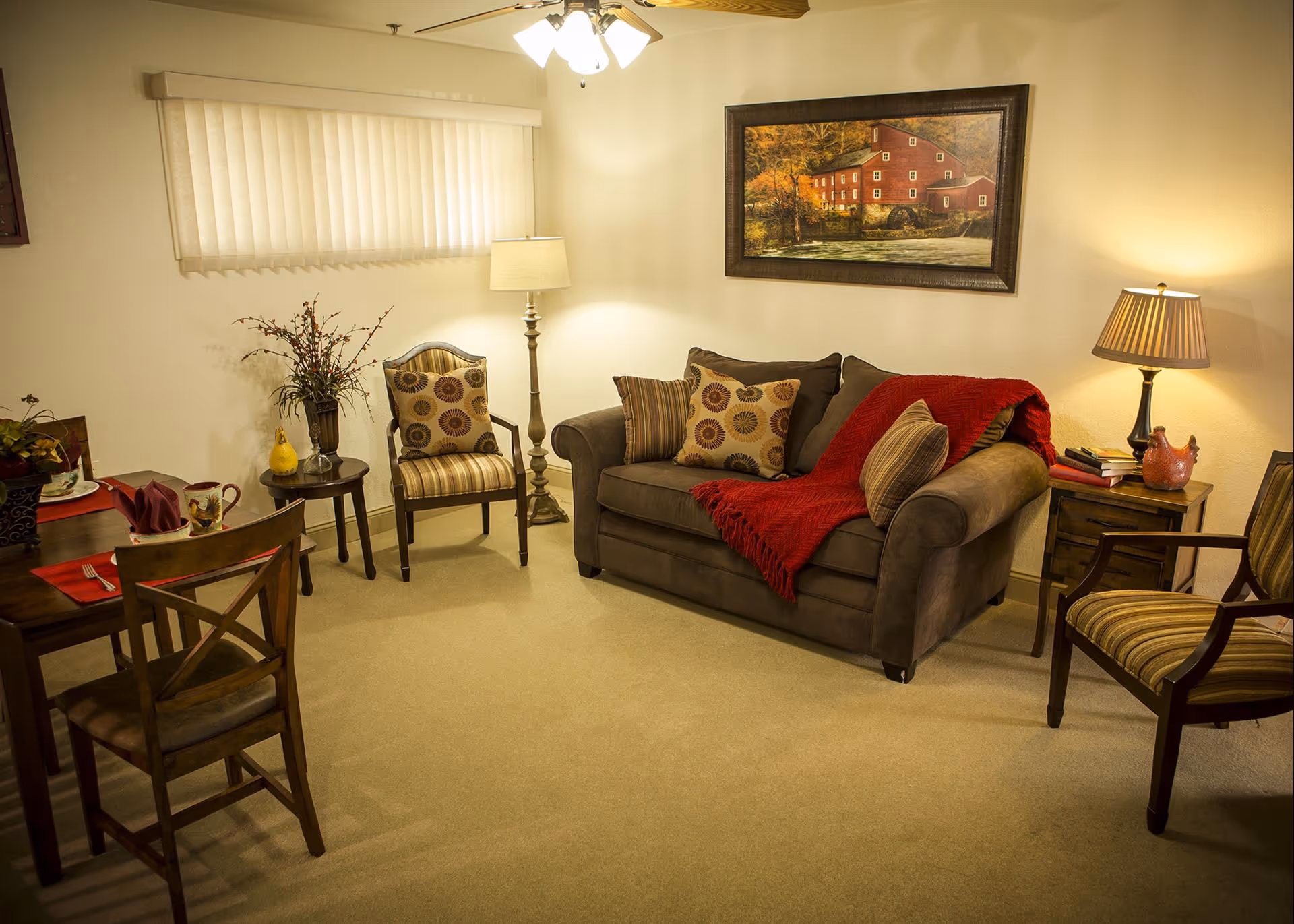 A cozy living room with a brown sofa adorned with patterned pillows and a red throw blanket. Two striped armchairs flank the sofa, and a wooden side table with a lamp and decorative items sits beside one chair. A floor lamp stands next to another armchair near a window with vertical blinds. A wooden dining table with chairs is partially visible on the left side, set with mugs and napkins. A framed painting of a red mill by a river hangs on the wall above the sofa.