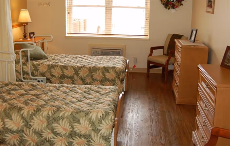 A room with two single beds covered with matching floral patterned bedspreads. There is a wooden dresser and a chair near the window with blinds. A lamp is on a small table next to one of the beds, and the floor is wooden.