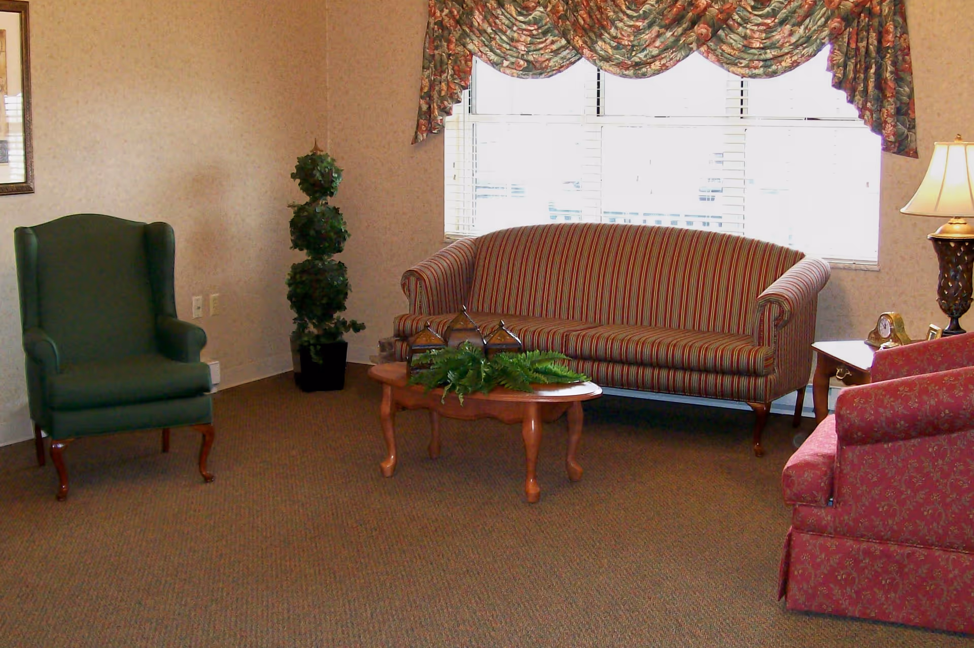 A cozy sitting area with a striped sofa, green and red armchairs, a wooden coffee table, lamp, and a large window with floral valance.