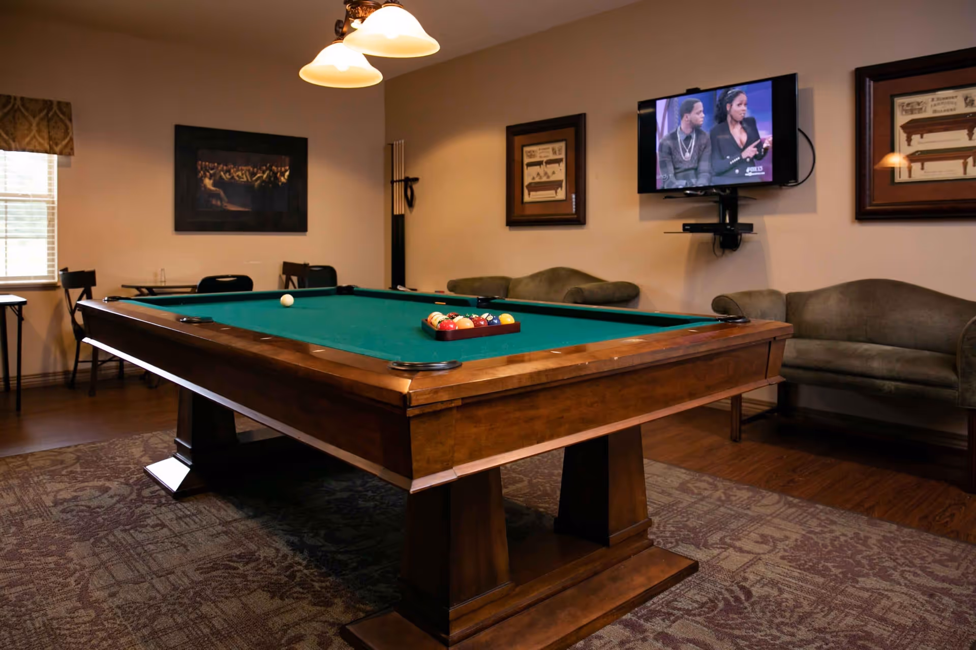 A cozy game room with a wooden pool table in the center, a rack of pool cues on the wall, a flat-screen TV mounted on the wall showing a talk show, two green upholstered sofas, framed pictures on the walls, and a window with a valance letting in natural light.