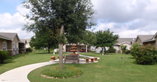 Outdoor view of Seguin Assisted Living facility showing a green lawn with a curved concrete pathway, a central tree surrounded by a circular flower bed, a multi-tiered water fountain with benches around it, and single-story residential buildings in the background under a partly cloudy sky.