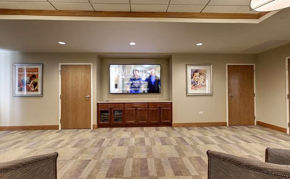 A spacious room with a large flat-screen TV mounted on the wall displaying Evergreen Senior Living information. Below the TV is a wooden cabinet with multiple compartments. The room has two closed wooden doors on either side of the TV, framed vintage-style posters on the walls, and patterned carpet flooring. The ceiling has recessed lighting and a wooden trim.