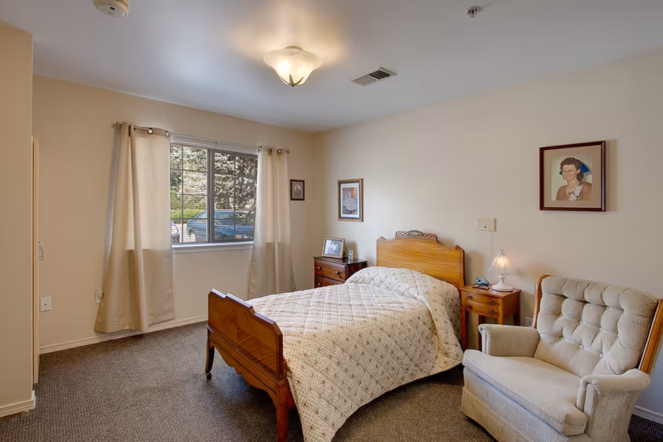 A cozy bedroom with a wooden bed covered with a patterned quilt, a cushioned armchair, two wooden nightstands with a lamp and framed photos, beige curtains on a window showing a parked car outside, and framed portraits on the walls.