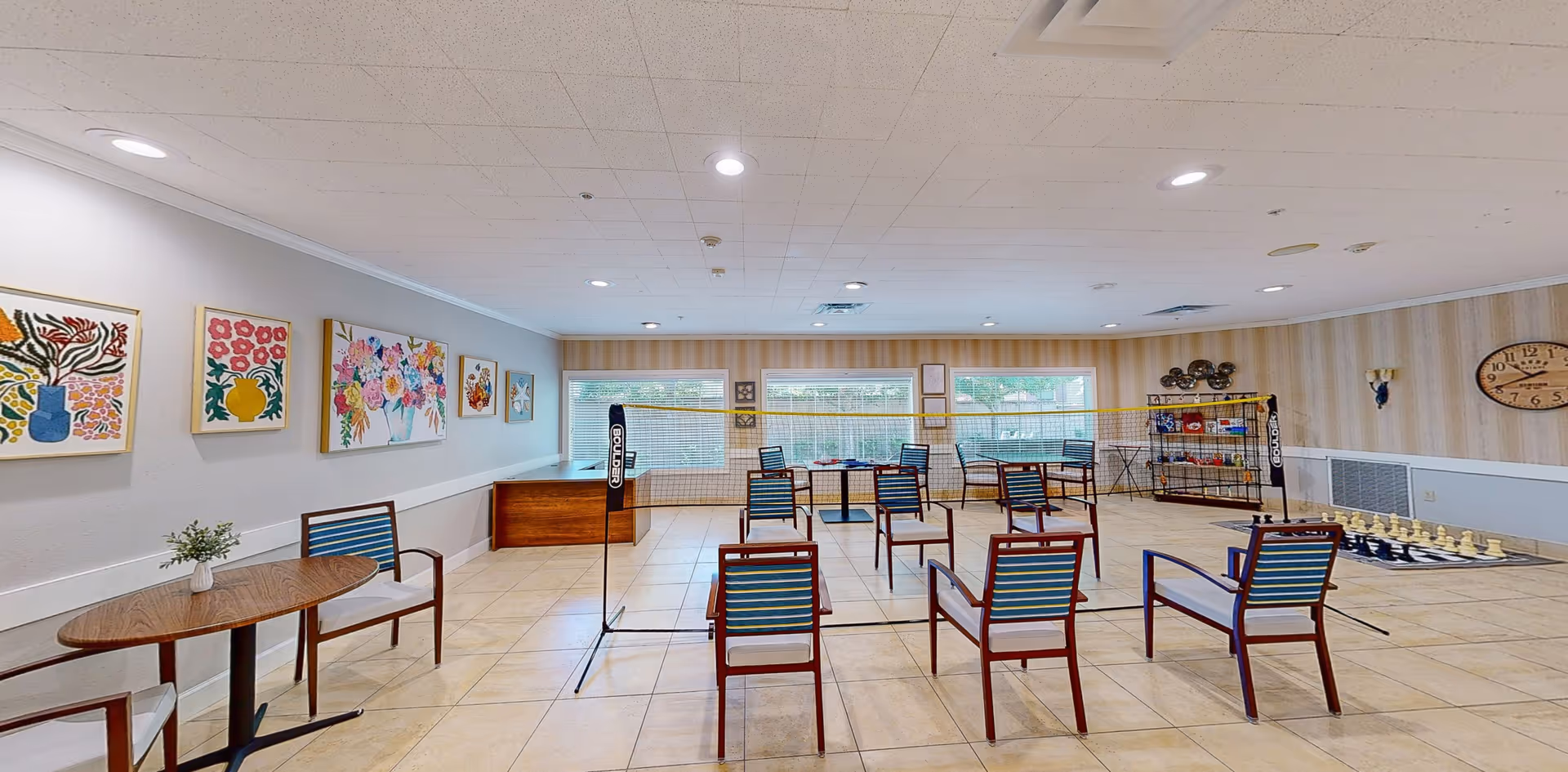 A spacious activity room with a tiled floor and beige walls decorated with colorful floral paintings. The room is set up for seated badminton with a net in the center and chairs arranged on both sides. There is a large chess set on the floor to the right and a shelving unit with games and supplies against the far wall. Large windows let in natural light, and a clock is mounted on the right wall.