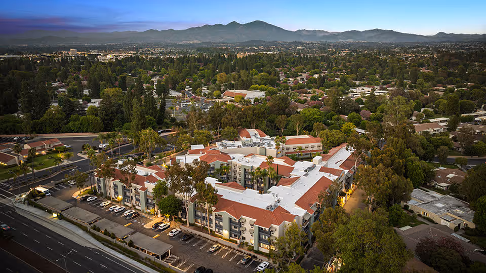 Aerial view of a large senior living facility with white and red roofs surrounded by trees and residential neighborhoods, with mountains visible in the background during dusk.
