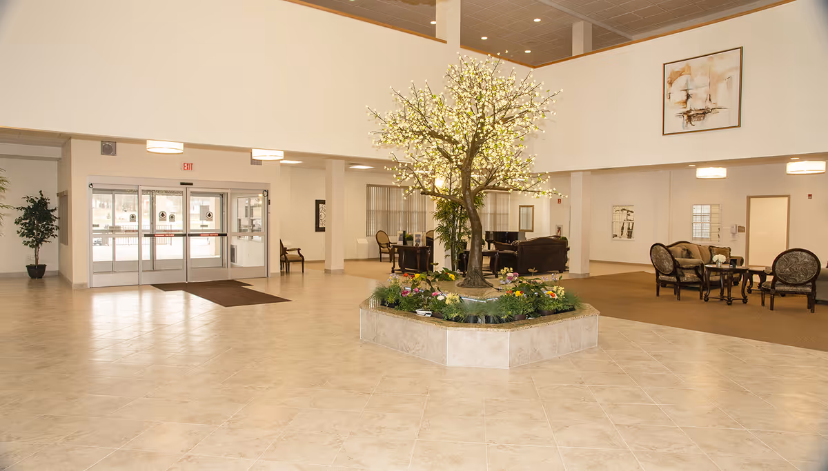 Spacious senior living facility lobby with a large artificial tree centerpiece surrounded by flowers. The area has tiled flooring, seating areas with chairs and tables, and large glass entrance doors. The walls are decorated with framed artwork.