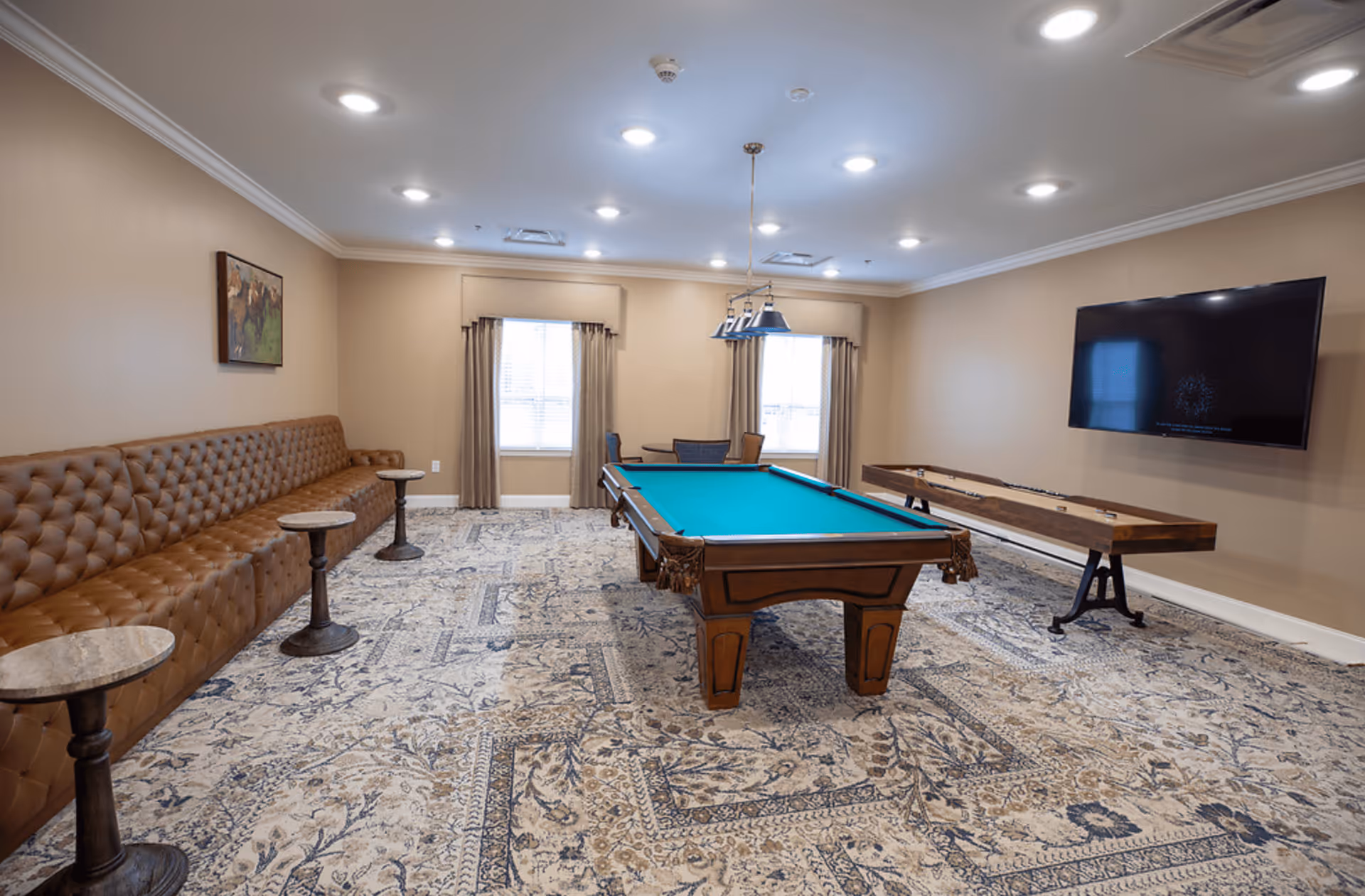 A recreational room with a pool table in the center, a long brown tufted leather bench along one wall with small round tables in front of it, a shuffleboard table against the opposite wall, a large flat-screen TV mounted on the wall, two windows with beige curtains, and a patterned carpet covering the floor.