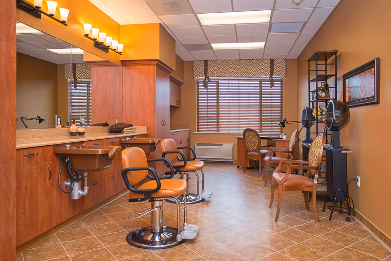 Interior of a salon area in a senior living facility with two brown salon chairs in front of sinks and mirrors on the left, and additional seating with hair dryers on the right. The room has warm brown walls, tiled floor, and a window with blinds and a valance at the back.