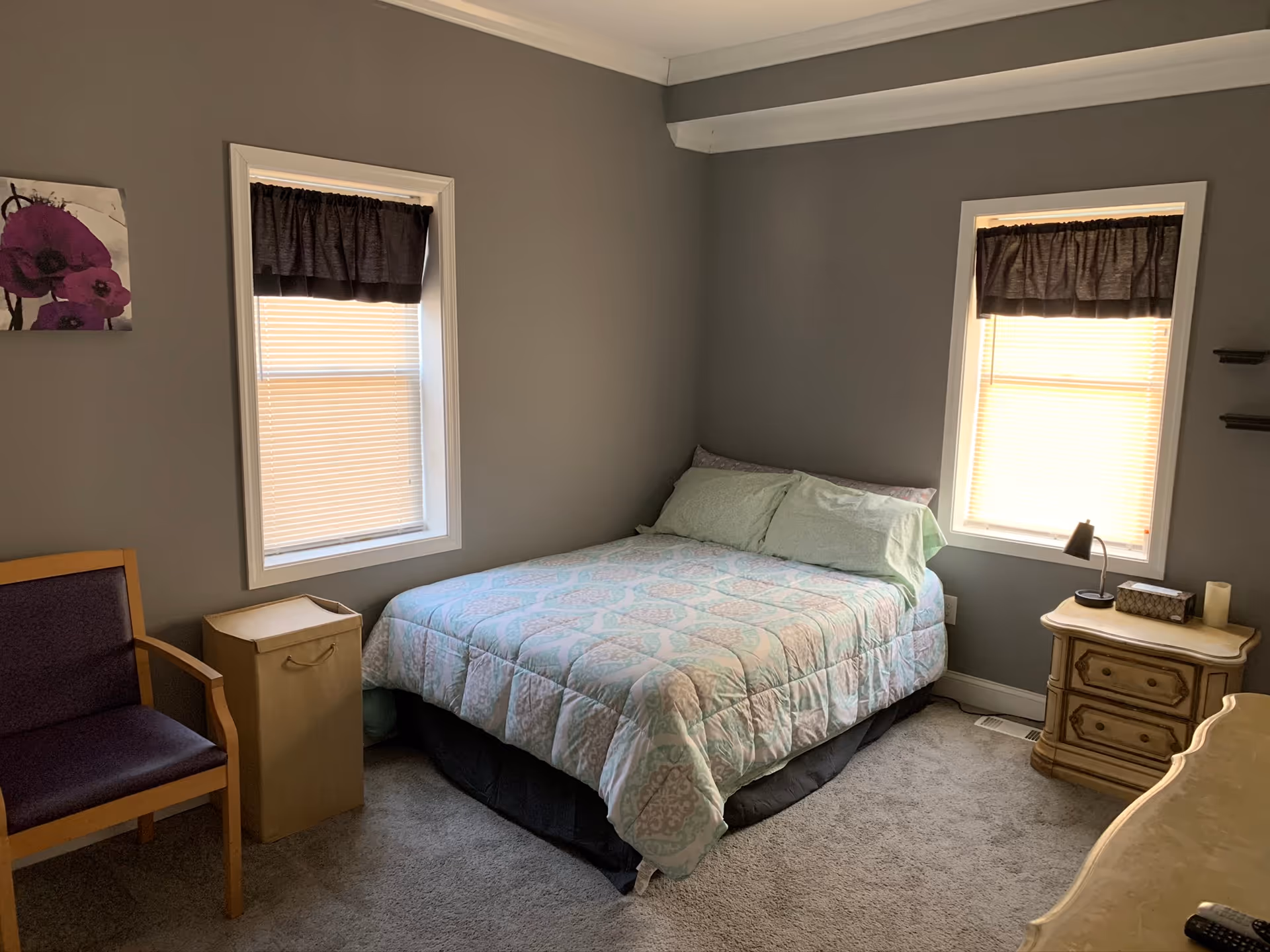 Small bedroom with a made double bed between two windows, a nightstand with lamp, chair and dresser.