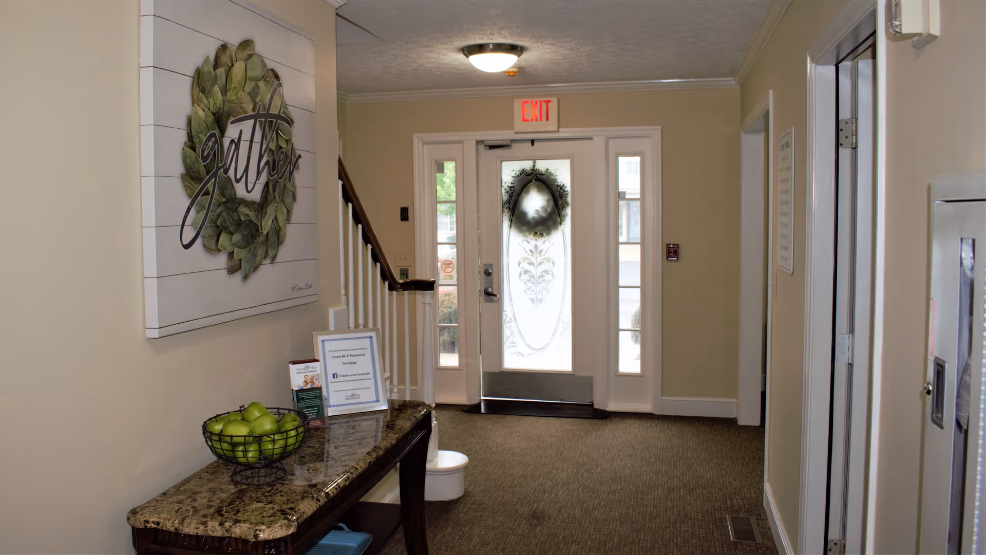 Interior hallway of an assisted living facility with a granite-topped table holding a basket of green apples and informational brochures. A decorative wall art with the word 'gather' hangs above the table. At the end of the hallway is a glass door with frosted floral designs and a wreath, with an illuminated exit sign above it.