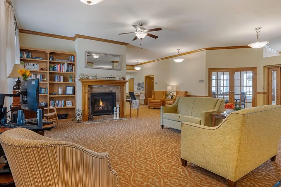 Spacious senior living common room with upholstered sofas and chairs, a fireplace, bookshelves, and a small computer workstation.