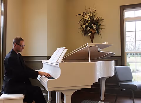 A man seated at a white grand piano playing in a bright communal room with chairs, windows, and a floral arrangement on the wall.