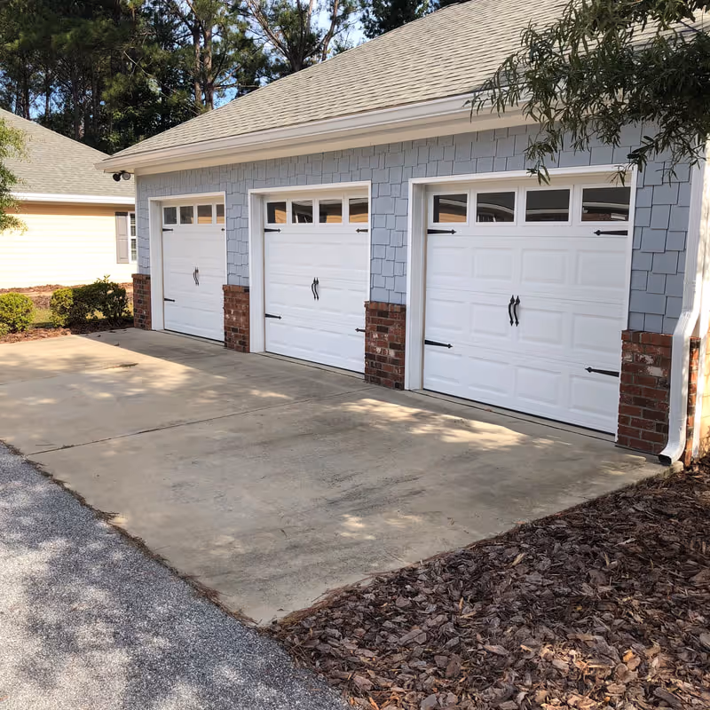 Exterior view of a building with three white garage doors, each with decorative black handles and hinges. The building has light blue siding with brick accents at the base and a gray shingled roof. There are trees and shrubs around the building and a concrete driveway in front.