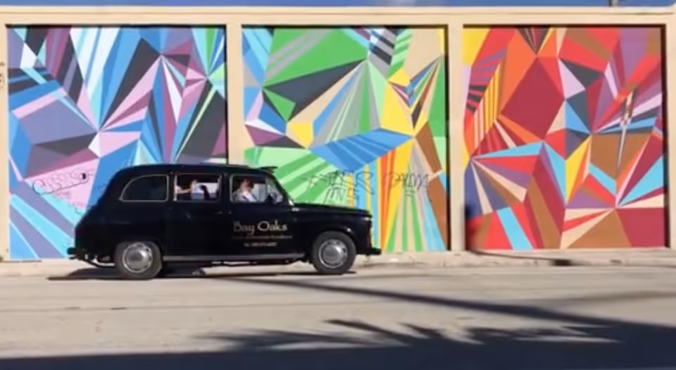 A black vintage car with 'Bay Oaks Historic Retirement Residence' written on the door is driving past a wall with three large colorful geometric murals in blue, green, and red tones.