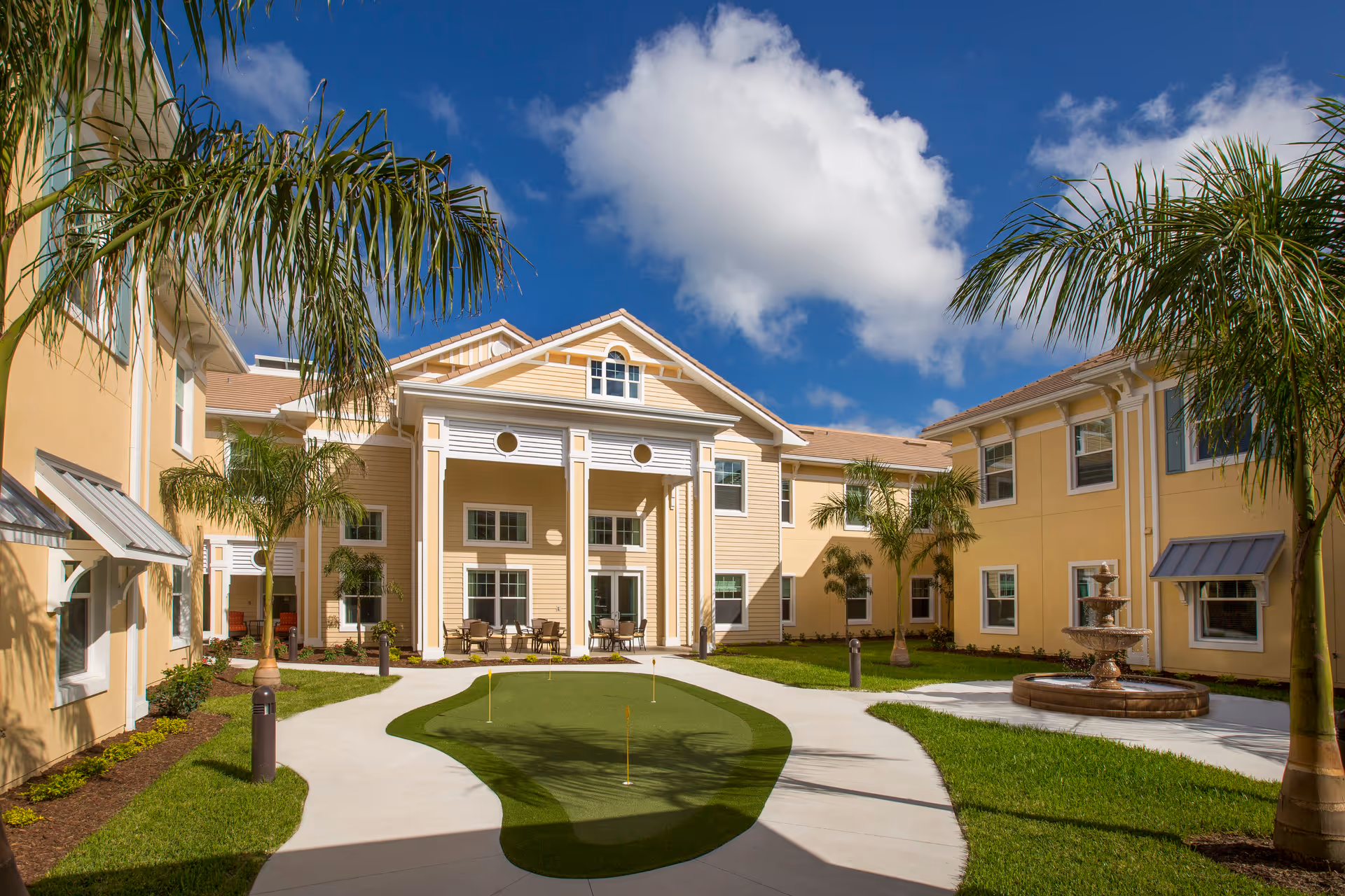 Outdoor courtyard area of a senior living facility with a putting green, palm trees, a fountain, and seating areas under a bright blue sky with a few clouds.