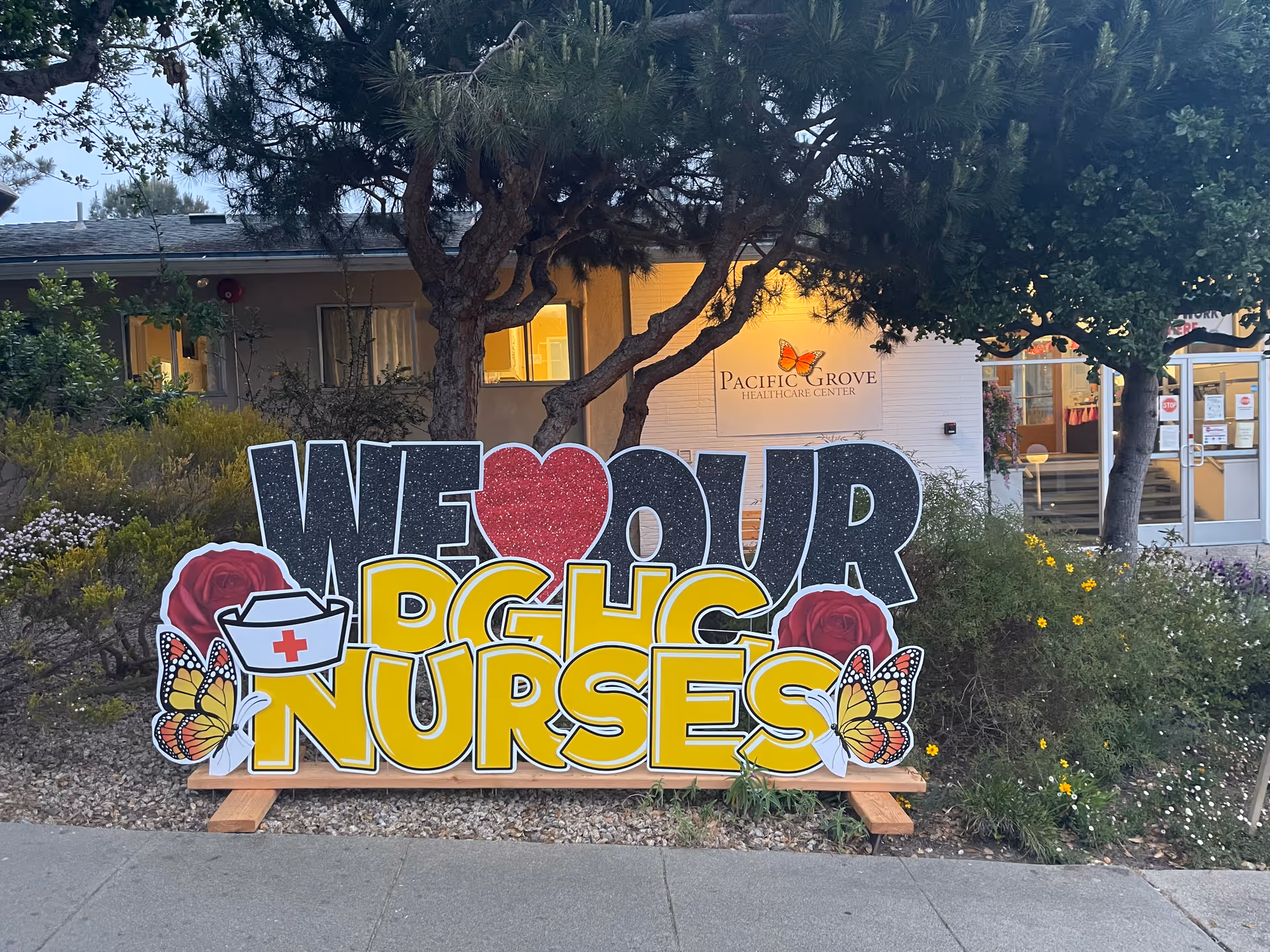 Outdoor view of Pacific Grove Healthcare Center with a large colorful sign in front that reads 'WE ♥ OUR PGHC NURSES' decorated with roses, butterflies, and a nurse's hat. The building entrance and windows are visible in the background surrounded by trees and bushes.