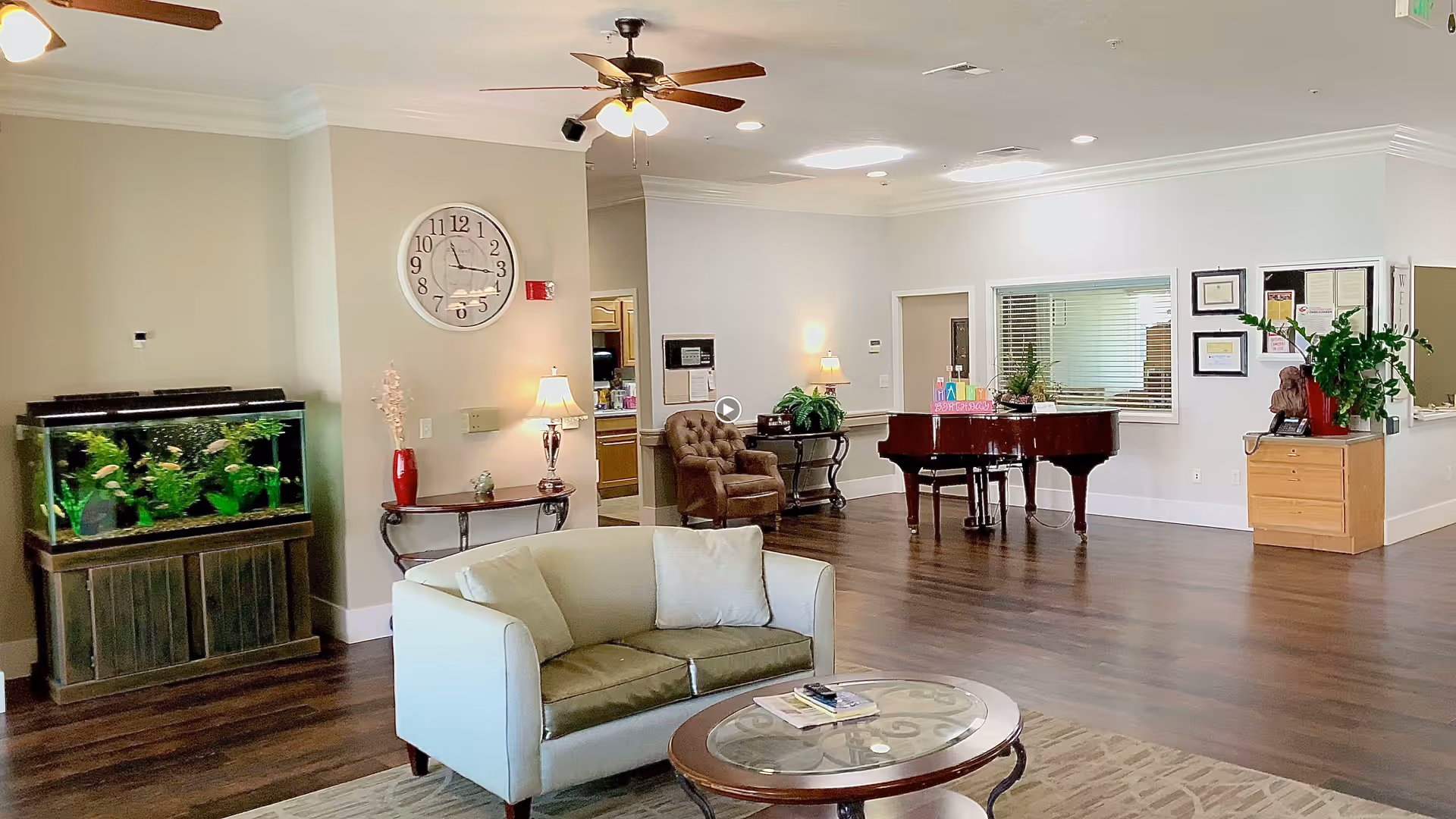 A spacious living room area in Ridgeview Gardens Assisted Living featuring a white loveseat with green cushions, a round glass coffee table, a large aquarium on a wooden stand, a wall clock, two table lamps on side tables, a brown armchair, a grand piano, and various plants and framed certificates on the walls.