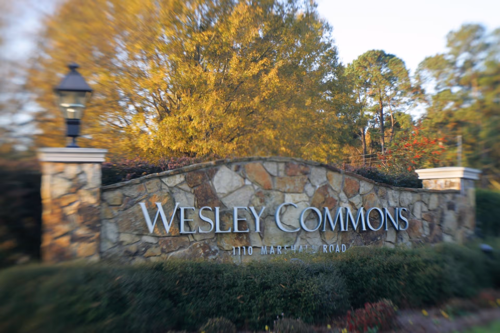Stone entrance sign for Wesley Commons with the address 1110 Marshall Road, surrounded by greenery and trees with autumn foliage.