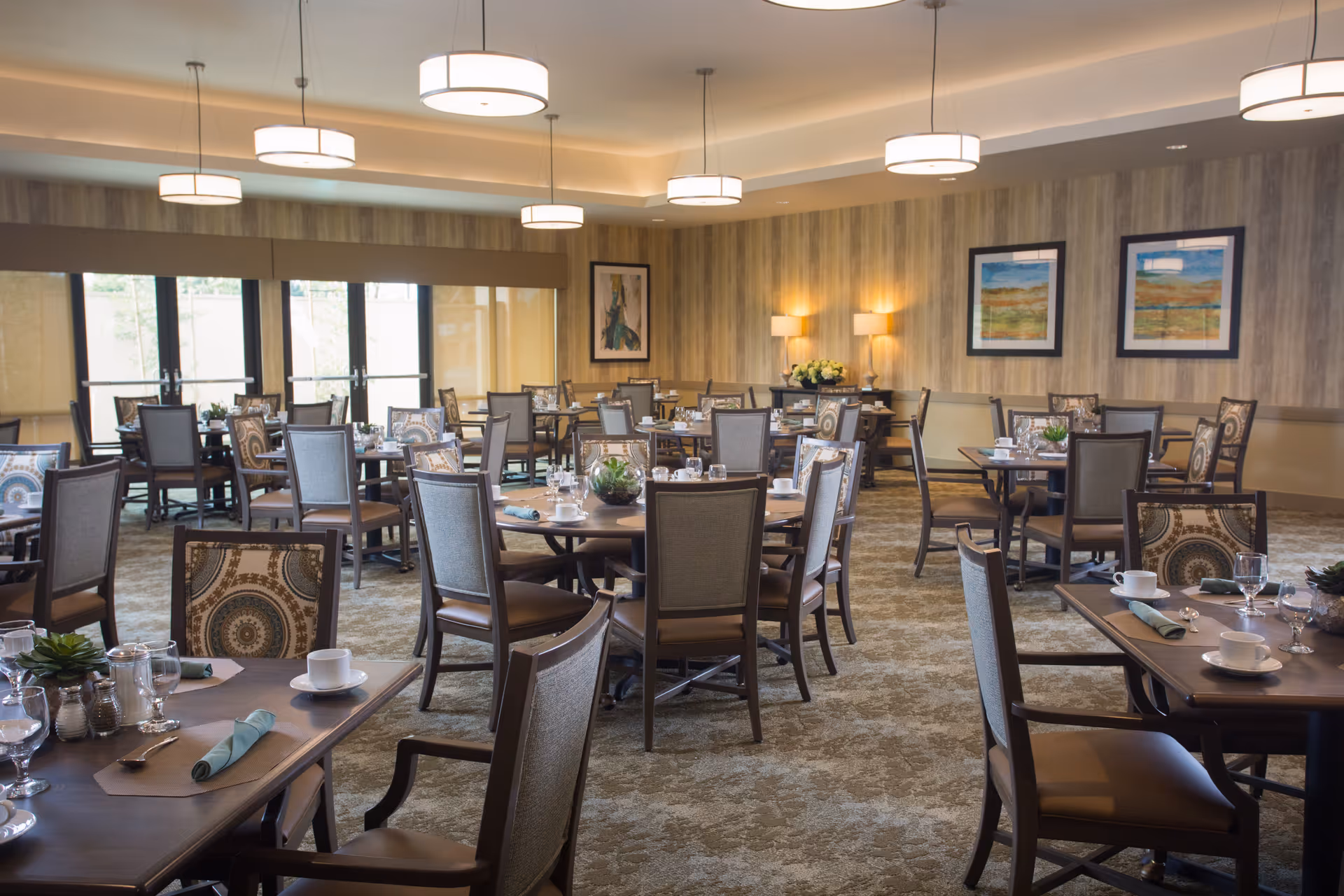 A spacious dining room with multiple round and rectangular tables set with cups, glasses, napkins, and silverware. The room features patterned chairs, carpeted flooring, large windows with blinds, modern ceiling lights, framed artwork on the walls, and soft wall lighting.