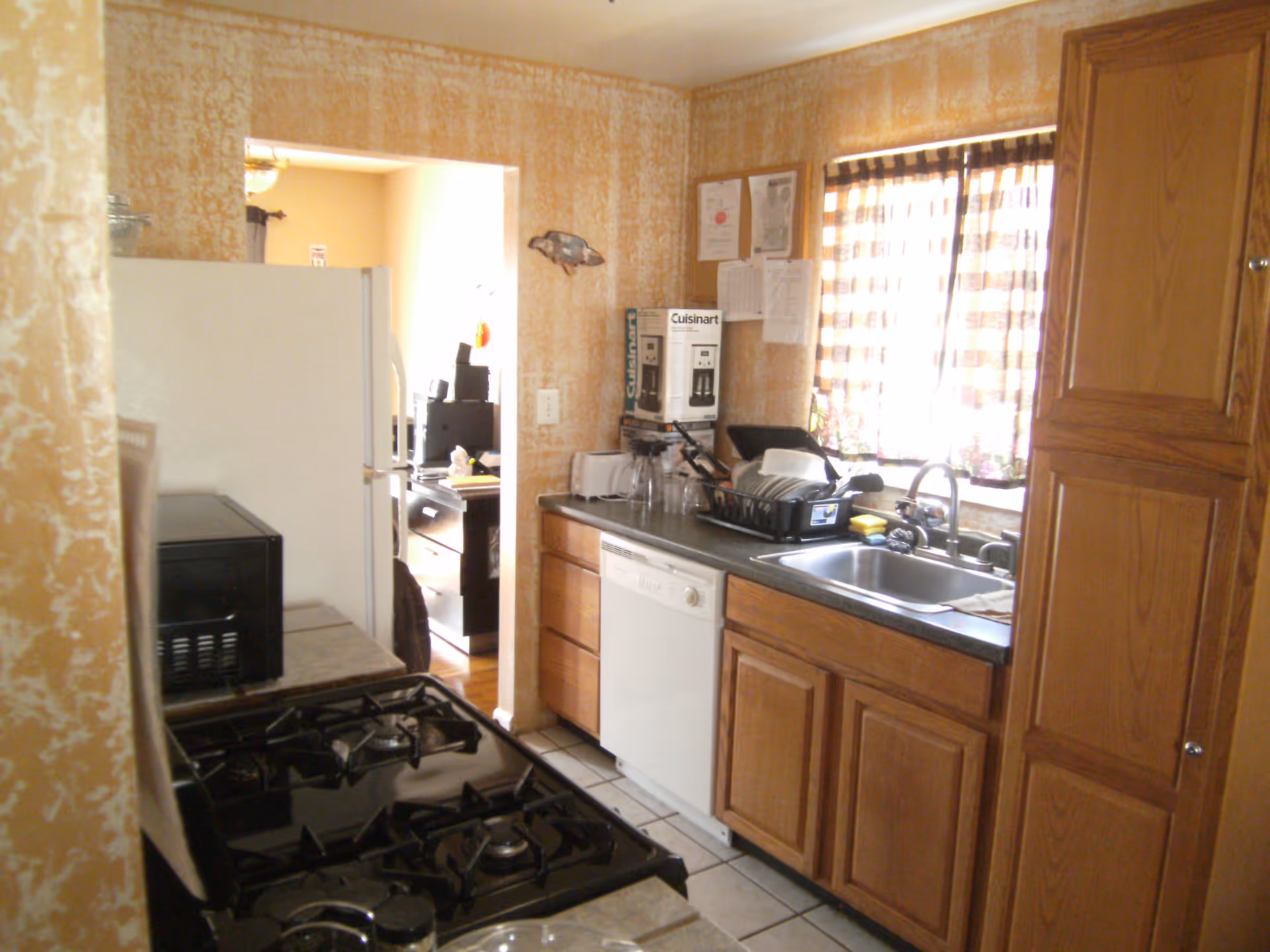 A small kitchen with wooden cabinets, a black gas stove, a white dishwasher, a stainless steel sink, and a window with checkered curtains. There are kitchen appliances and dishes on the counter, and a refrigerator is visible through an open doorway leading to another room.