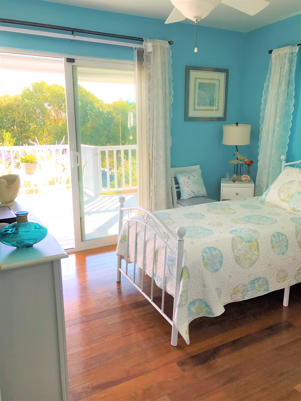 A bright bedroom with turquoise walls and wooden flooring featuring a white metal bed frame with a quilted bedspread in pastel colors. There is a white nightstand with a lamp and decorative items, a chair with a cushion, and a dresser with a blue glass vase. Sheer white curtains frame a sliding glass door that opens to a balcony with a white railing and greenery outside.