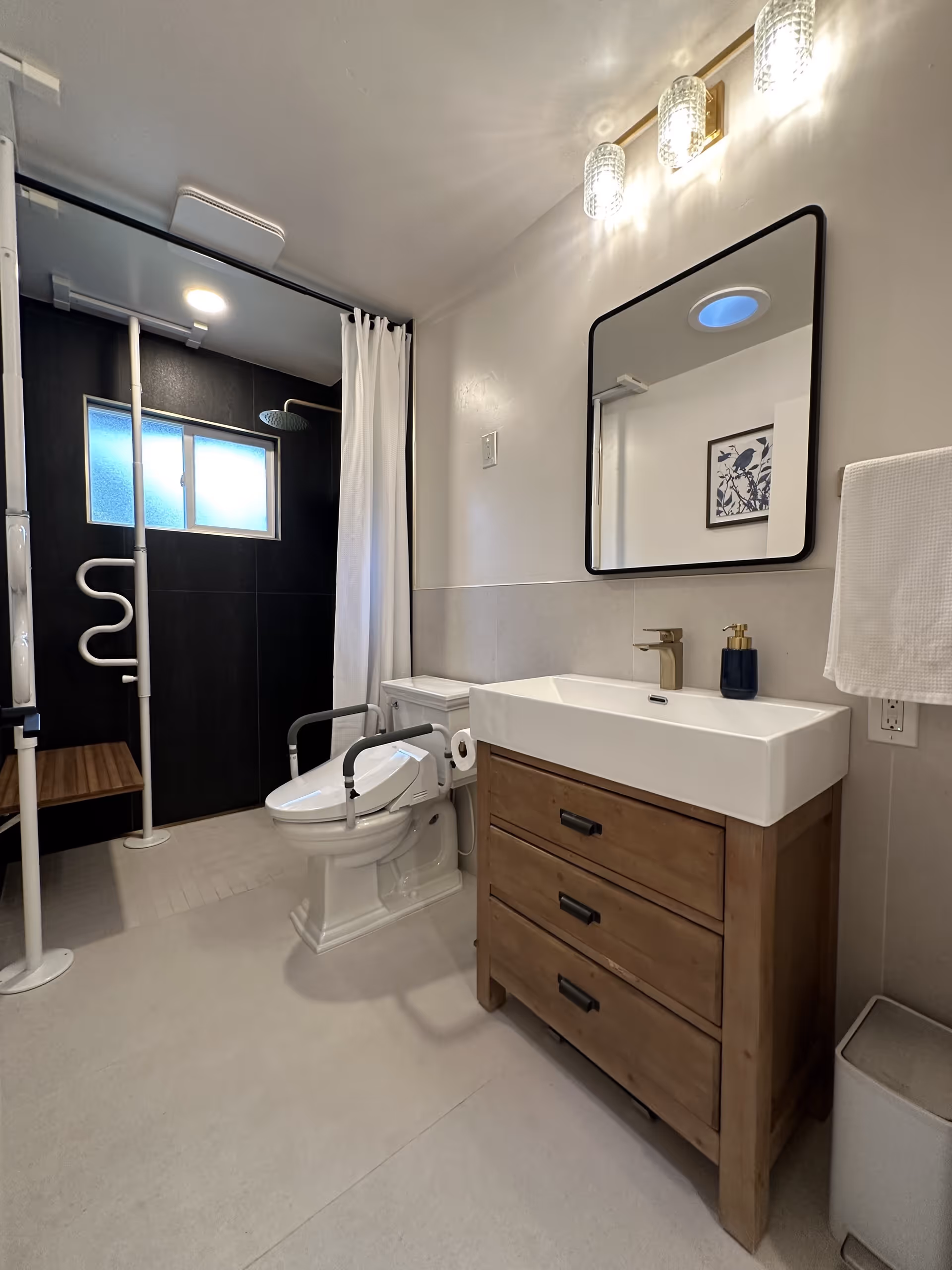 A modern bathroom with a white toilet equipped with safety rails, a wooden vanity with a white sink and gold faucet, a rectangular mirror with rounded corners above the sink, and a walk-in shower area with a white curtain and a wooden bench. The shower has black tiled walls and a frosted window. There are two wall-mounted lights above the mirror and a towel hanging on the right side.