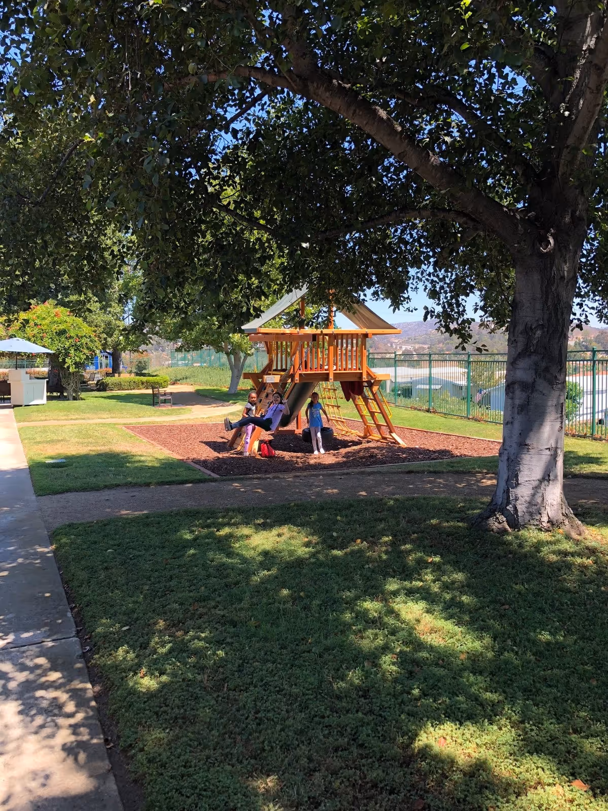 A sunny outdoor playground area with a wooden play structure featuring a slide, swings, and a climbing ladder. Three children are playing on the swings under the shade of a large tree. The area is surrounded by green grass, a paved walkway, and a metal fence in the background with hills visible beyond.