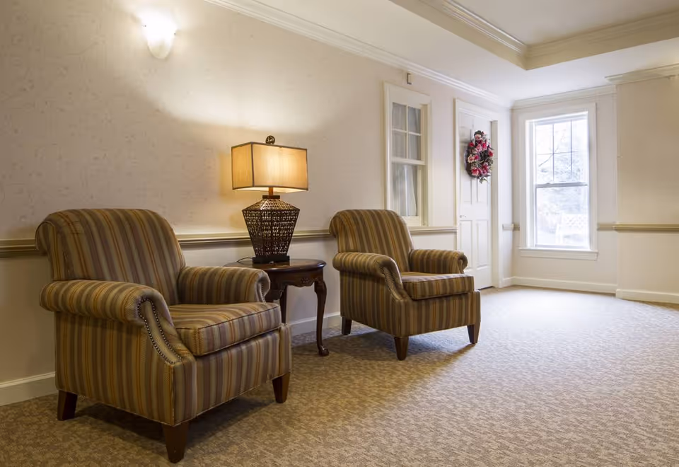 A cozy interior seating area with two striped armchairs positioned on either side of a small wooden side table with a decorative lamp. The room has beige patterned carpet, light-colored walls with subtle floral wallpaper, a door adorned with a floral wreath, and a large window letting in natural light.