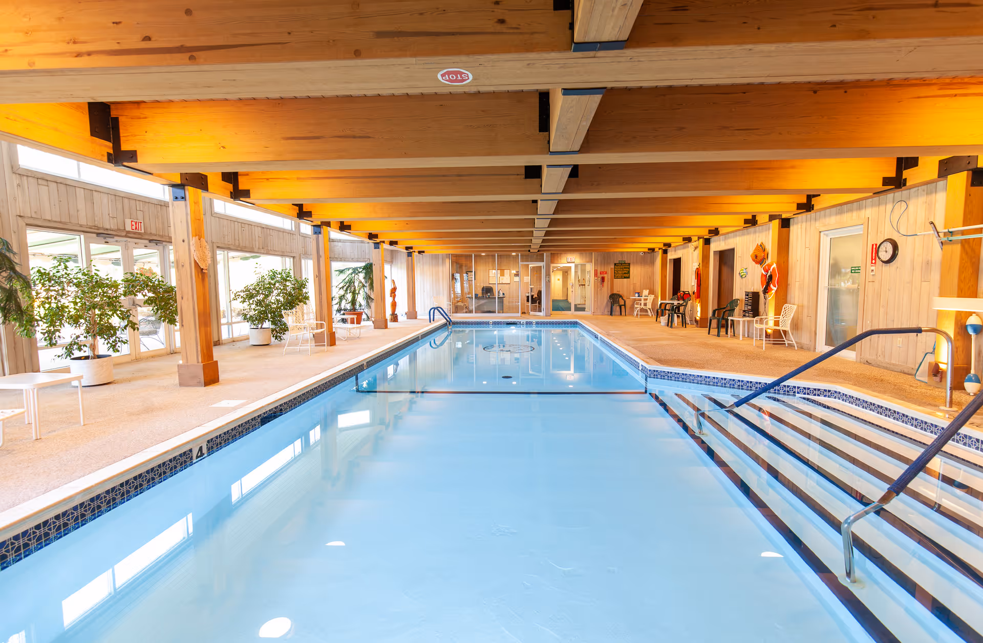 Indoor swimming pool area with clear blue water, wooden ceiling beams, potted plants along the sides, chairs and tables placed around the pool deck, and large windows letting in natural light.