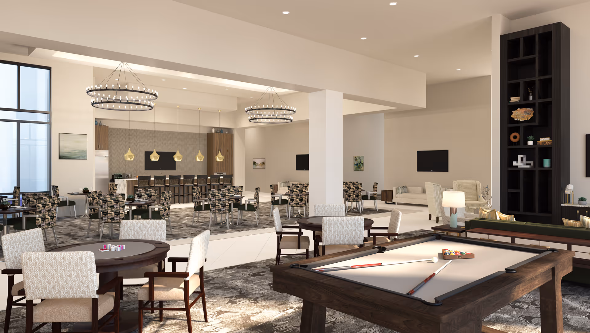 A spacious and modern common area in a senior living facility featuring a pool table with cues and balls, a round card table with chairs, multiple seating areas with patterned chairs and sofas, two large circular chandeliers, and a kitchen area with a bar and stools in the background.