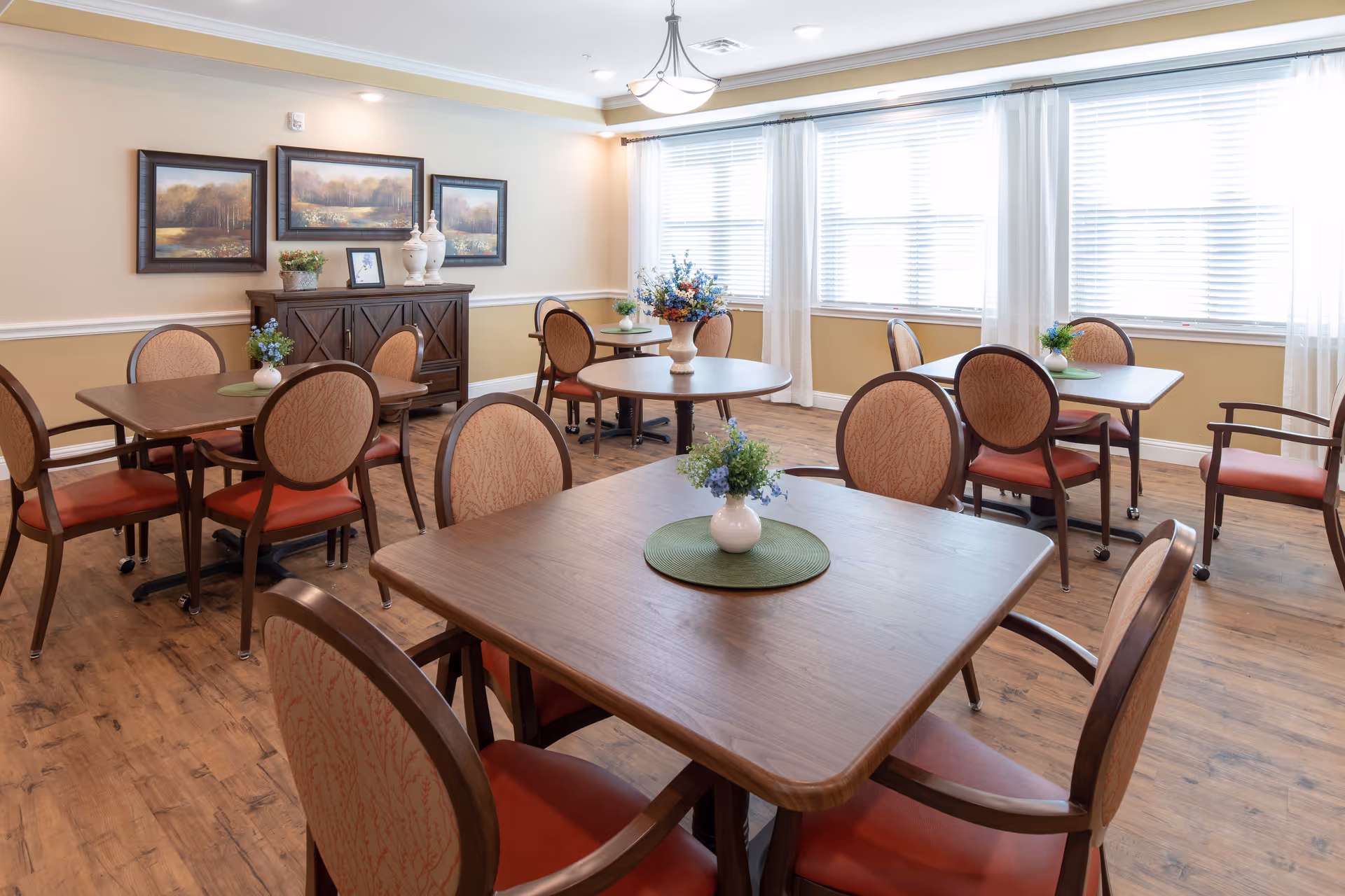 A bright dining room with multiple wooden tables and cushioned chairs arranged neatly. Each table has a small vase with flowers on a green placemat. The room has large windows with white blinds and sheer curtains, wooden flooring, and a sideboard against the wall with framed landscape paintings and decorative items on top.