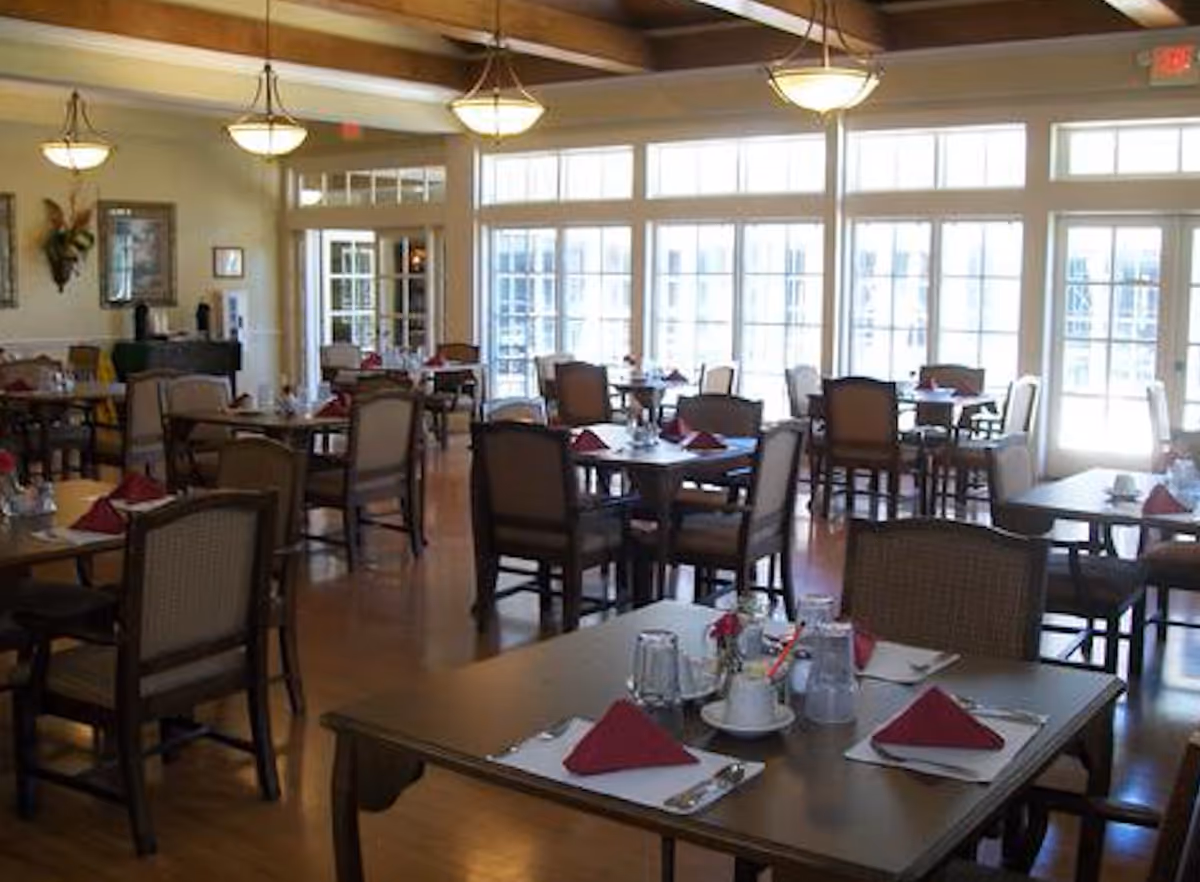 A bright dining room with multiple tables set with red napkins, glasses, and silverware. The room has large windows letting in natural light, wooden floors, and hanging light fixtures. Chairs are arranged around the tables, and there are decorative wall hangings and plants on the walls.