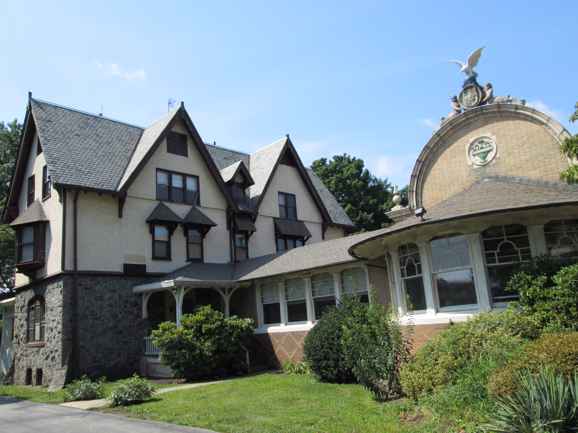 Exterior view of a large, historic-style building with a stone foundation and beige upper walls, featuring multiple gabled roofs and decorative architectural details. The building is surrounded by green shrubs and a well-maintained lawn under a clear blue sky.
