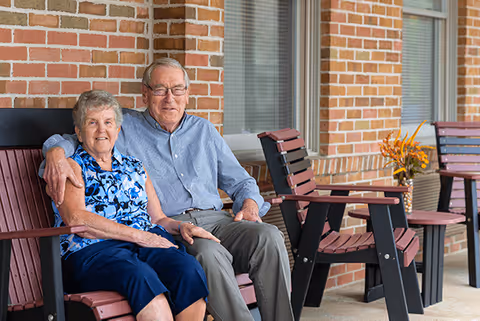 An elderly couple sitting closely together on a wooden bench outside a brick building, smiling at the camera. There are additional benches and a small table with a vase of flowers nearby.