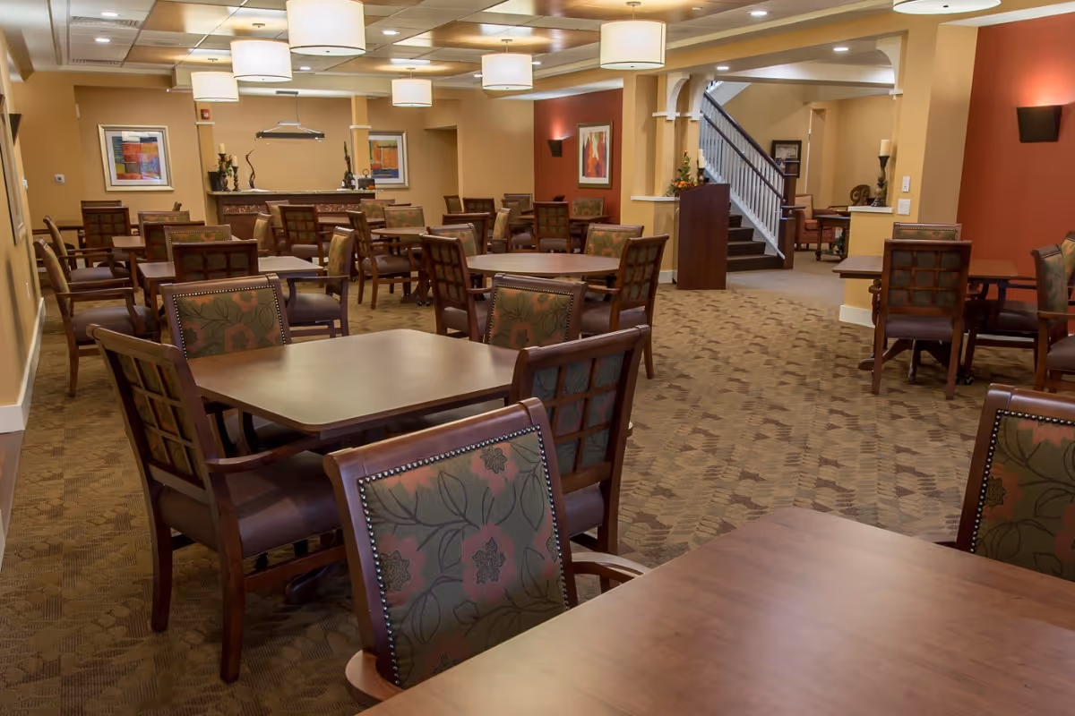 A spacious dining room with multiple wooden tables and chairs featuring floral patterned upholstery. The room has warm beige and red walls, carpeted floors, ceiling lights, and framed artwork on the walls. A staircase is visible in the background.