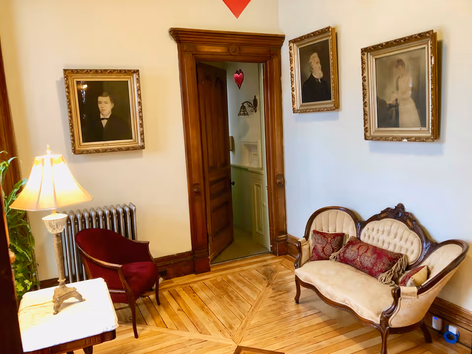 Small vintage sitting room with an upholstered settee and armchair, framed portraits, wooden doorway and parquet floor.