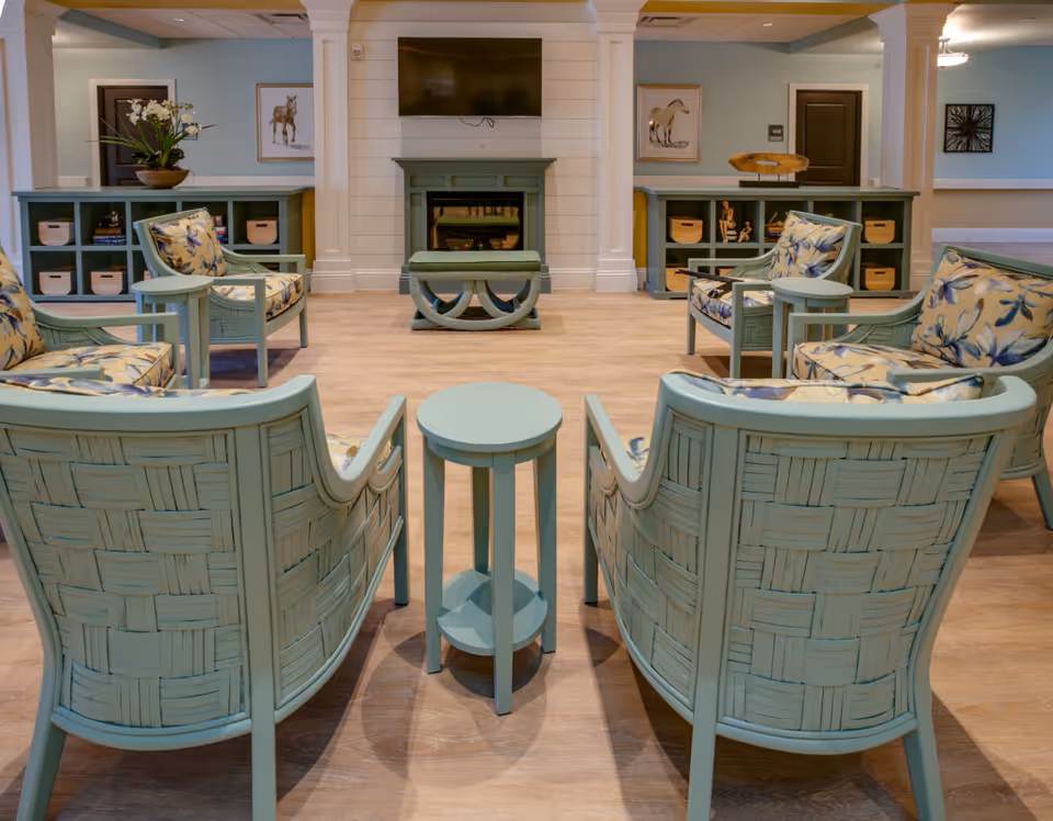 A cozy living room area with light blue woven armchairs featuring floral cushions arranged around small round tables. The room has light wood flooring, a fireplace with a mounted TV above it, and built-in shelving units with decorative items and baskets. The walls are painted light blue with white trim and columns.