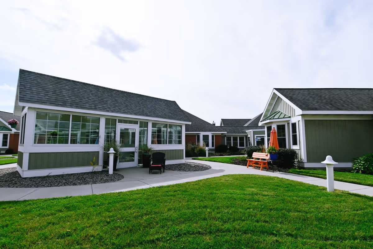 Exterior view of a senior living facility with green lawns, paved walkways, and multiple single-story buildings with green and white siding and dark shingled roofs. There is a bench with an orange umbrella and some potted plants near one of the buildings.