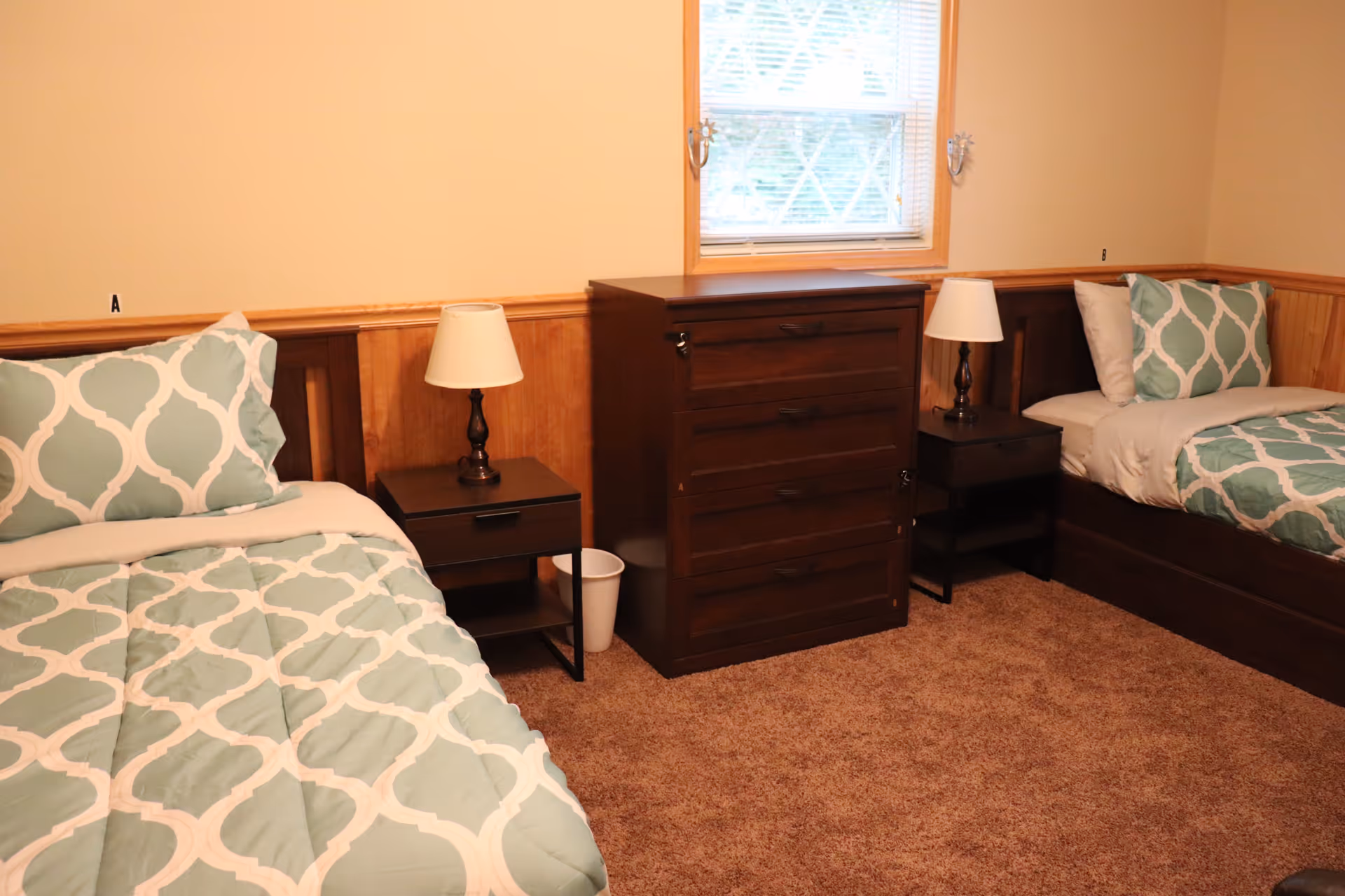 A bedroom with two twin beds, each with a green and white patterned comforter and matching pillows. Between the beds are two dark wooden nightstands with white lamps. A dark wooden chest of drawers is placed between the nightstands under a window with blinds. The room has beige walls with wooden wainscoting and brown carpeted floor.