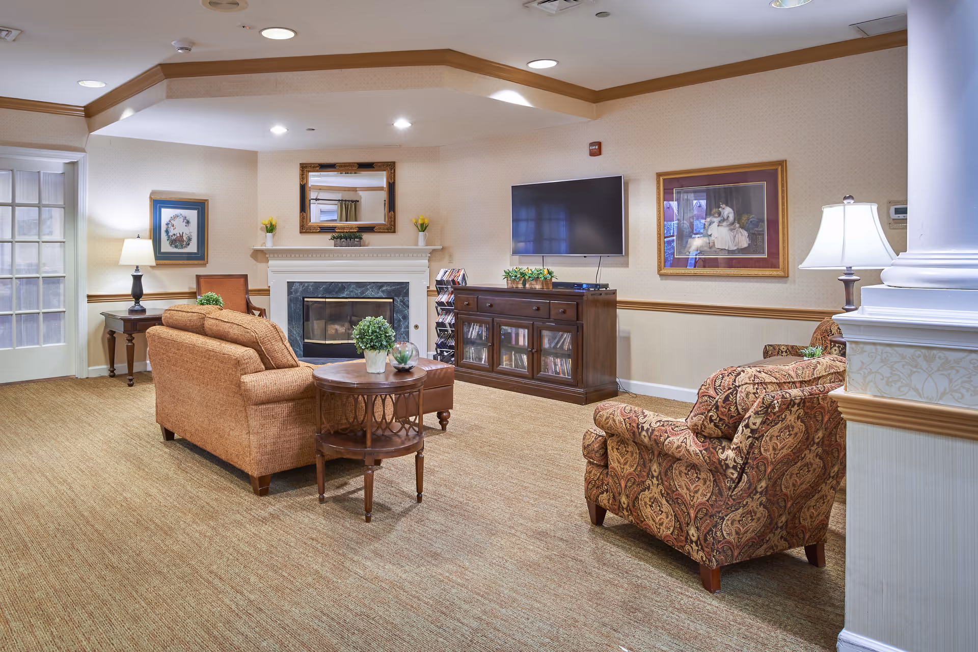 A cozy living room area with a beige couch and a patterned armchair facing a fireplace and a wall-mounted flat-screen TV. The room has carpeted floors, a wooden side table with a small plant, framed artwork on the walls, and warm lighting from table lamps.