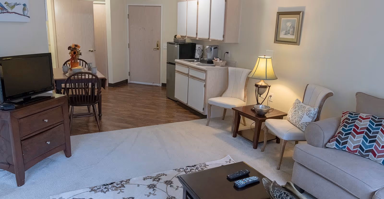 A cozy living area in a senior living facility featuring a beige sofa with colorful patterned pillows, two white cushioned chairs with a wooden side table and lamp between them, a wooden coffee table with remote controls, a small dining table with chairs, a TV on a wooden stand, and a kitchenette with cabinets, a mini fridge, and a coffee maker. The room has wood flooring near the entrance and carpet in the living area.