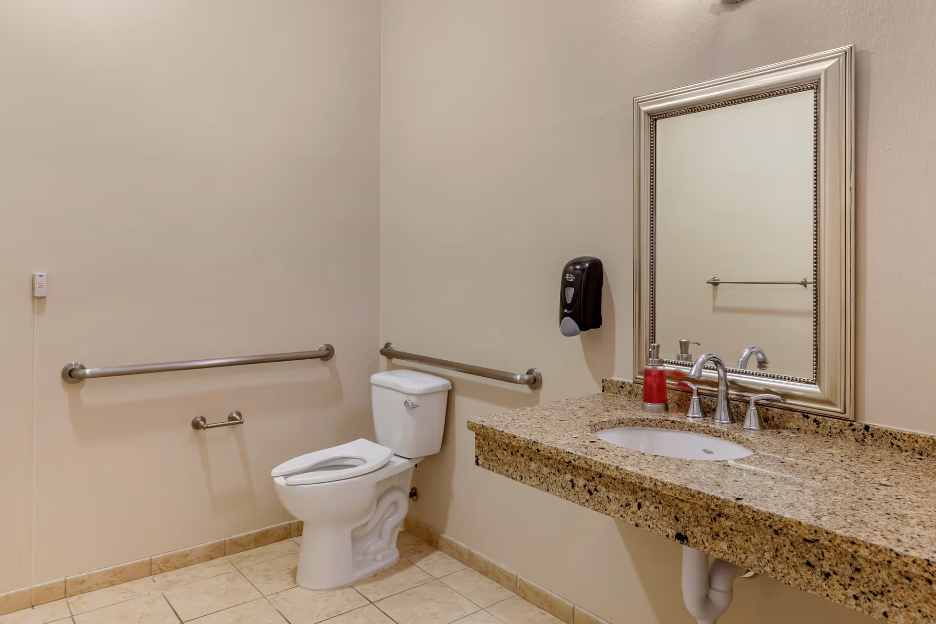 A clean and accessible bathroom with beige walls and tiled floor. The room features a white toilet with stainless steel grab bars on the walls around it. There is a granite countertop with an under-mount sink, a silver faucet, a soap dispenser with red liquid, and a large framed mirror above the sink. A black soap dispenser is mounted on the wall next to the mirror.