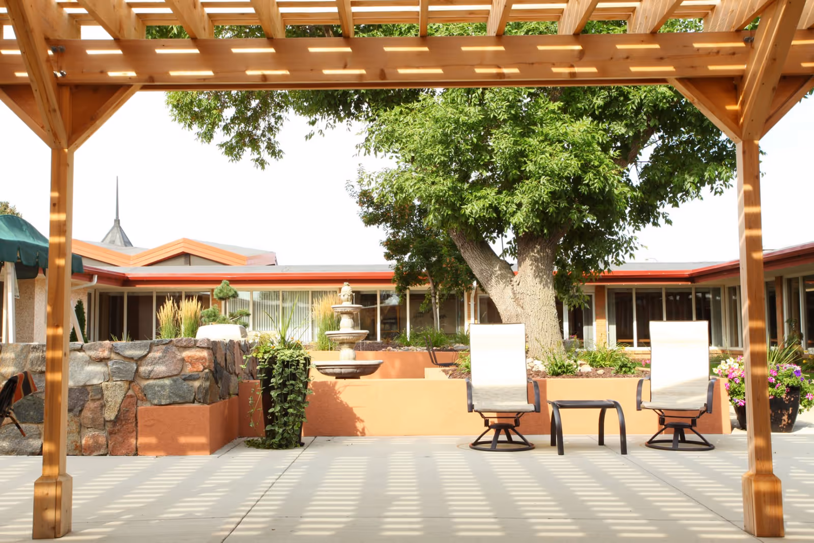Courtyard with a wooden pergola, two patio chairs and a small table, a fountain, and a large tree in front of the facility building.